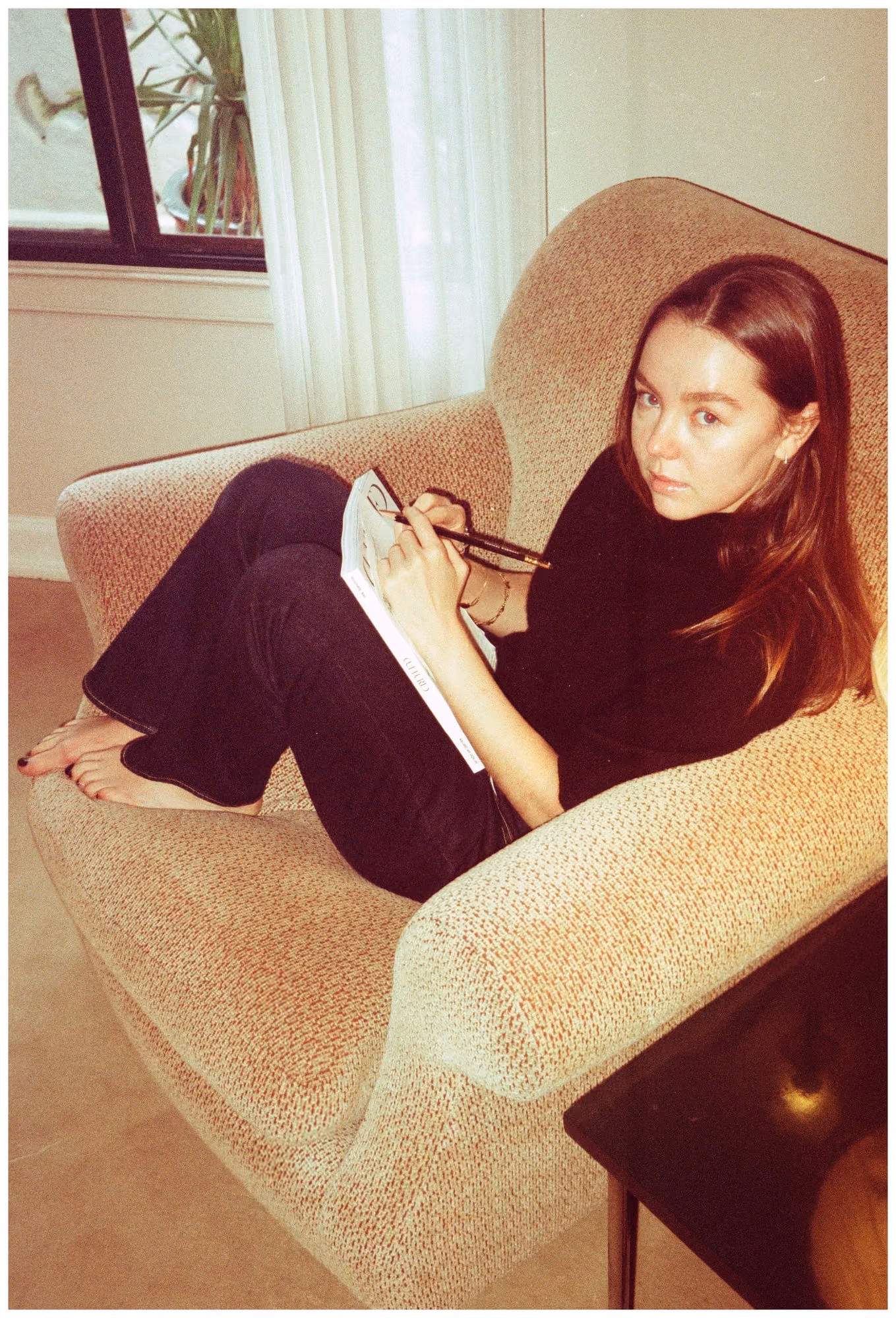 Alex Hanover reclining in a pale armchair with a notebook and pen, looking towards the camera, a window and houseplant behind her.